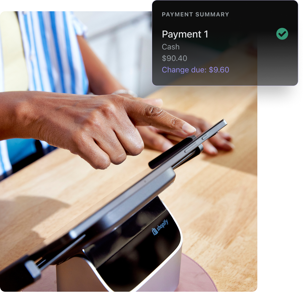 A closeup of a checkout counter where an associate where a blue and white striped overshirt is typing on a POS tablet. The tablet and stand are on a pale wood counter. Overlaid on the image is a Shopify POS UI screen indicating a payment summary.